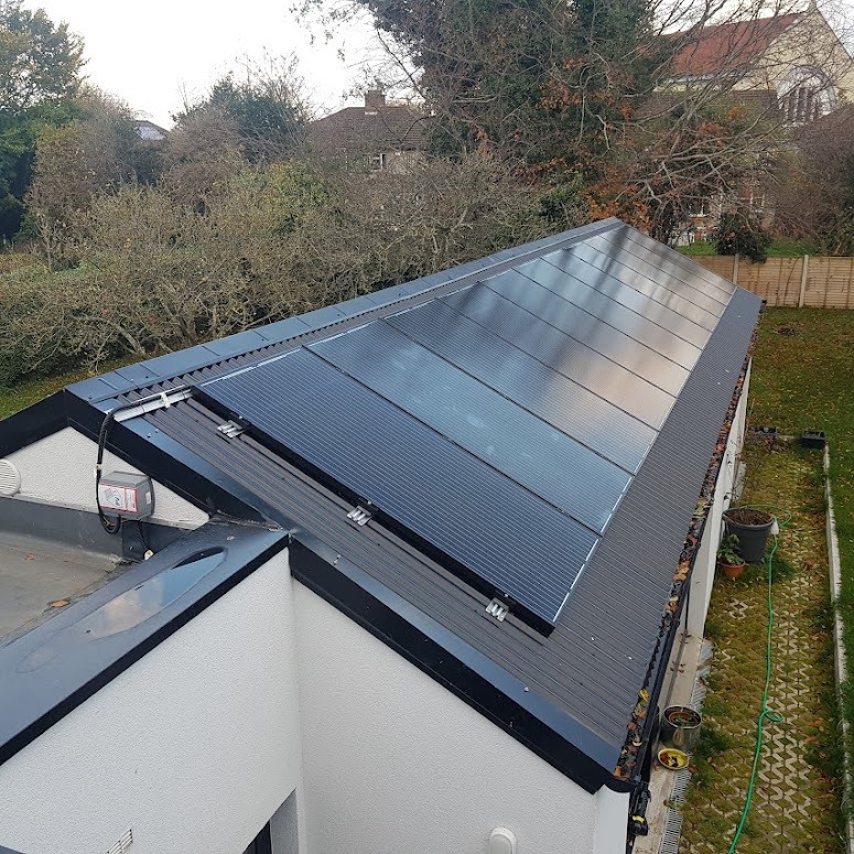 Panele fotowoltaiczne zamontowane na dachu budynku z blachy trapezowej, widok z góry na instalację i otaczający ogród z drzewami.