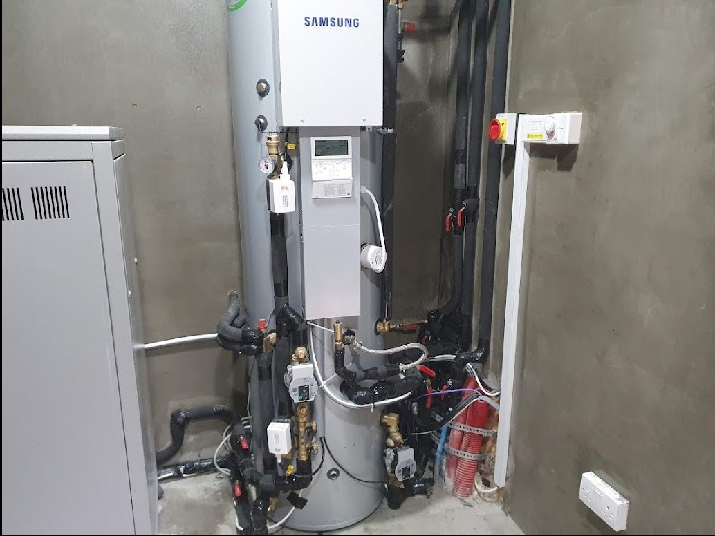 Szara, pionowa pompa ciepła Samsung z czarnymi rurami i przewodami w surowym, betonowym pomieszczeniu technicznym.