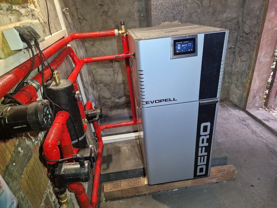 Nowoczesny piec Defro Evopell z wyświetlaczem, podłączony do instalacji grzewczej z czerwonymi rurami w kotłowni o surowym wykończeniu.