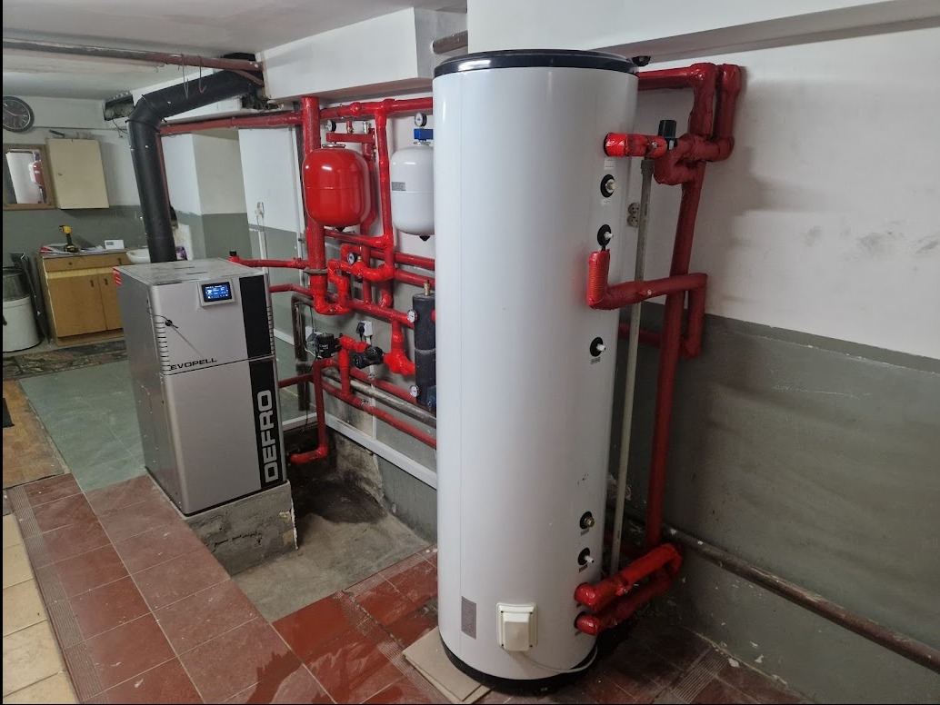 Kotłownia z kotłem na pellet Defro Evopell i białym zasobnikiem ciepłej wody, rury w kolorze czerwonym, płytki podłogowe w odcieniach brązu, widoczne elementy instalacji grzewczej.