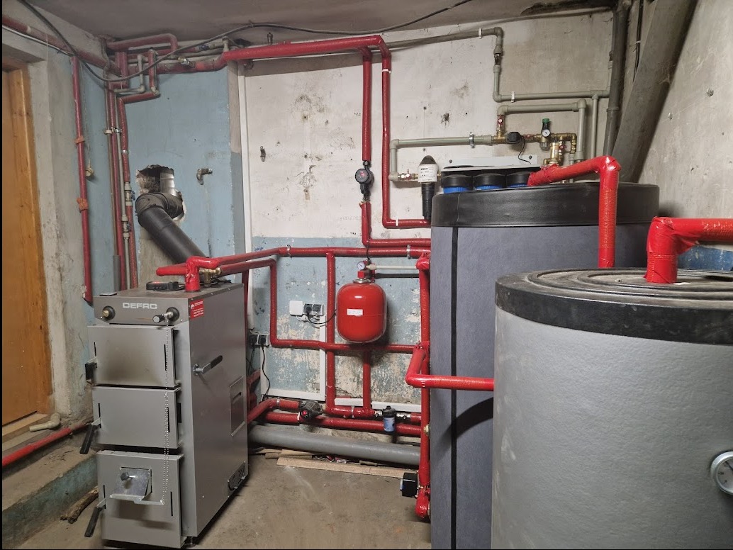 Szary piec Defro z podłączeniem do instalacji grzewczej z czerwonymi rurami i dwoma szarymi zbiornikami w piwnicy z betonowymi ścianami.