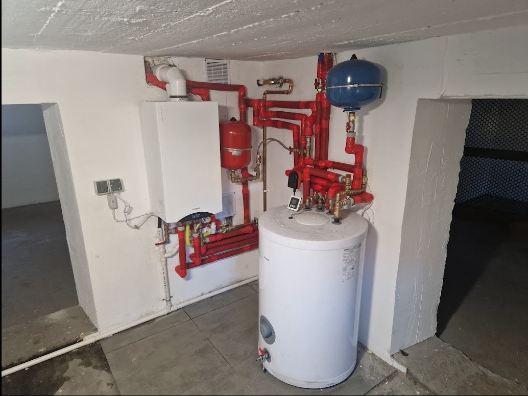 Kocioł gazowy kondensacyjny z podgrzewaczem wody i ekspansomatem, rury w kolorze czerwonym, instalacja w piwnicy z surowym betonowym stropem.