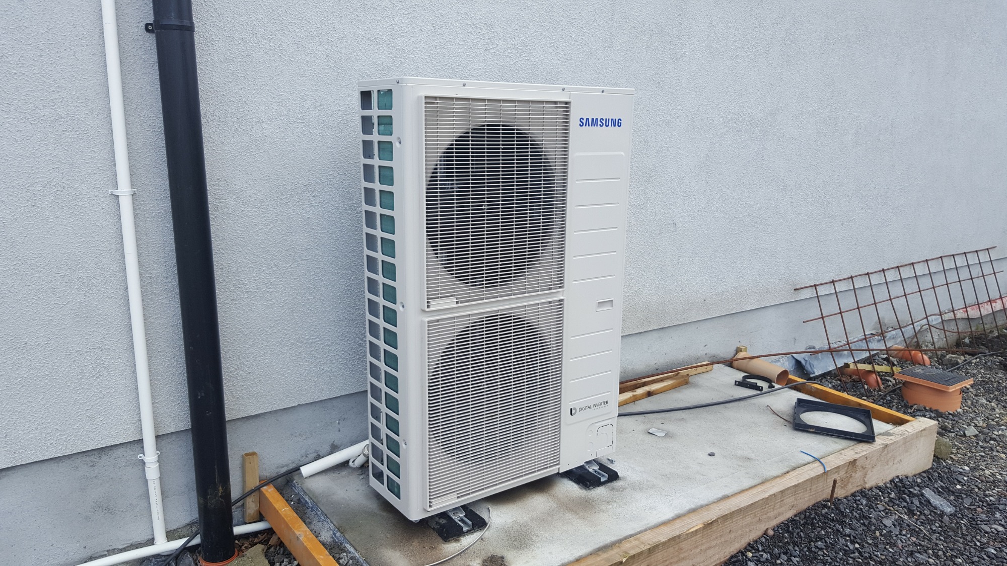 Biała jednostka zewnętrzna klimatyzacji Samsung zamontowana na betonowej podstawie przy ścianie budynku z widocznymi rurami odprowadzającymi skropliny, w tle ziemia i elementy konstrukcyjne.