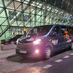 Niebieski bus firmy transportowej Rapid.net z logo i informacją o transferach na lotniska Pyrzowice i Okęcie, zaparkowany przed nowoczesnym budynkiem lotniska wieczorem.