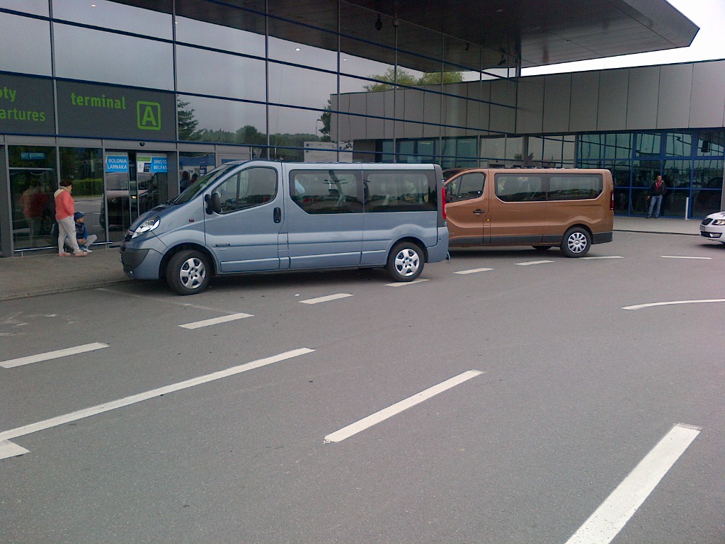 Dwa busy, jeden niebieski i jeden brązowy, zaparkowane przed wejściem do terminalu lotniska.