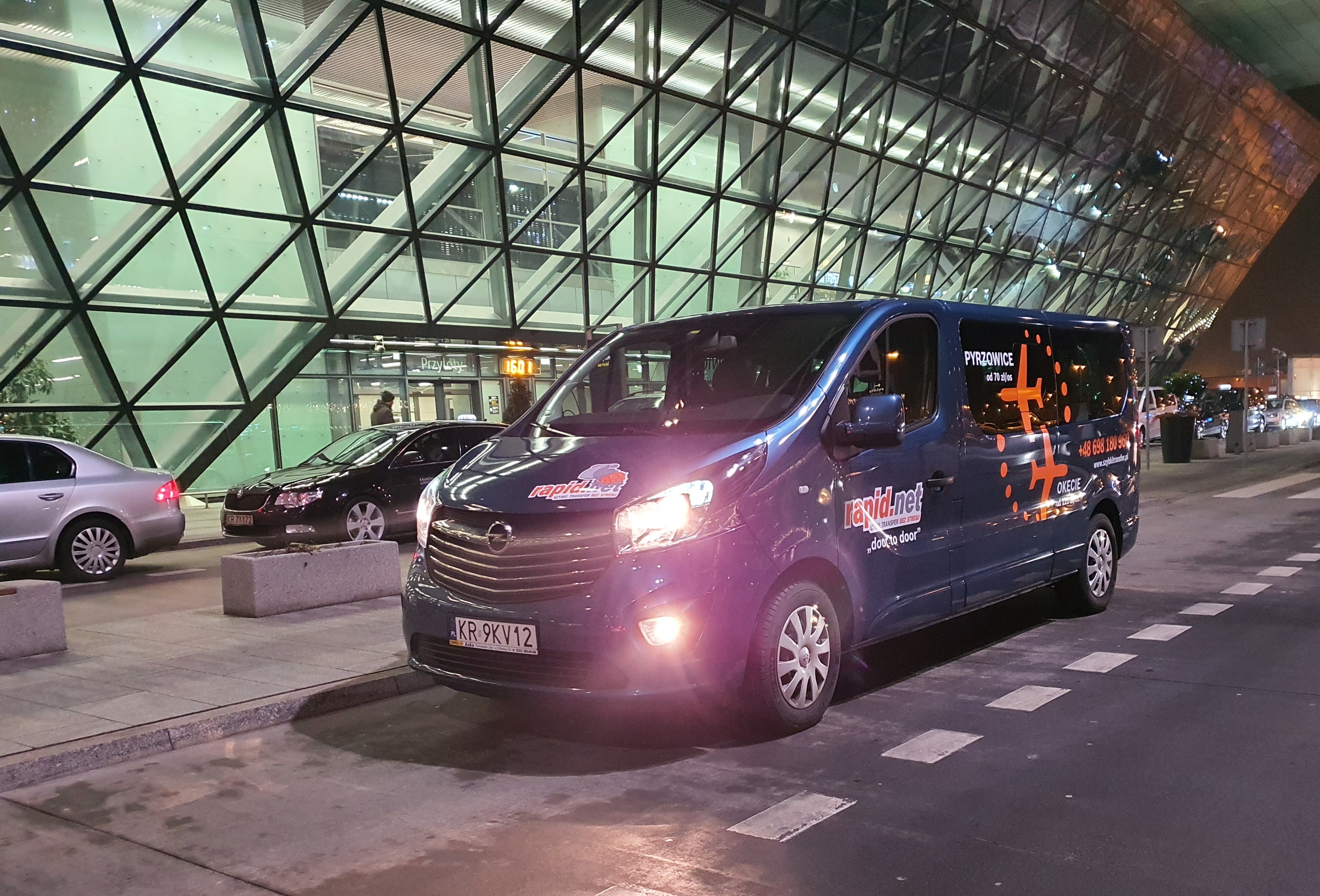 Niebieski bus firmy transportowej Rapid.net z logo i informacją o transferach na lotniska Pyrzowice i Okęcie, zaparkowany przed nowoczesnym budynkiem lotniska wieczorem.