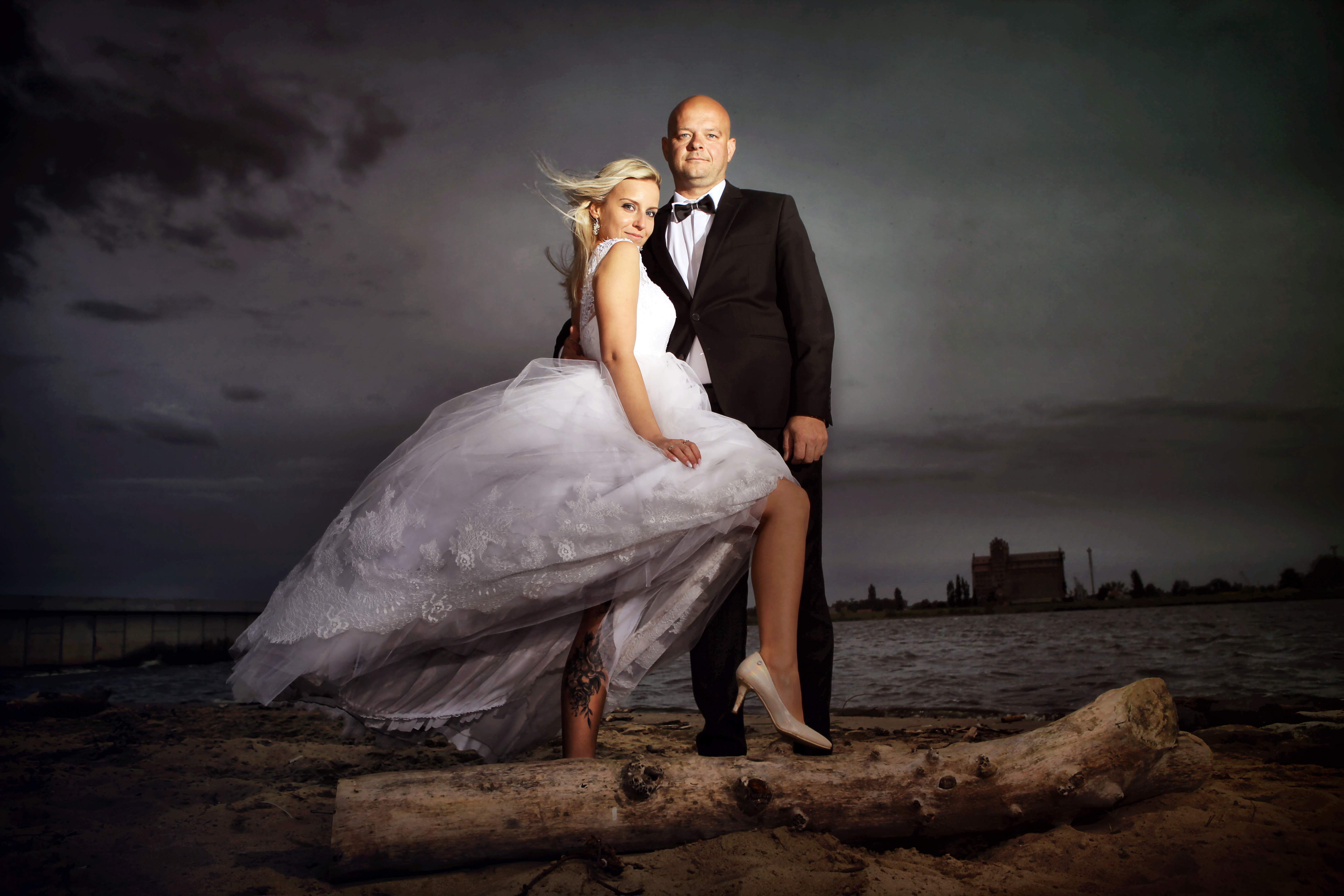 Elegancka para młoda pozuje na plaży na tle pochmurnego nieba i odległego budynku; panna młoda w białej sukni z falującą spódnicą i szpilkach opiera nogę na pniu drzewa, a pan młody w czarnym...