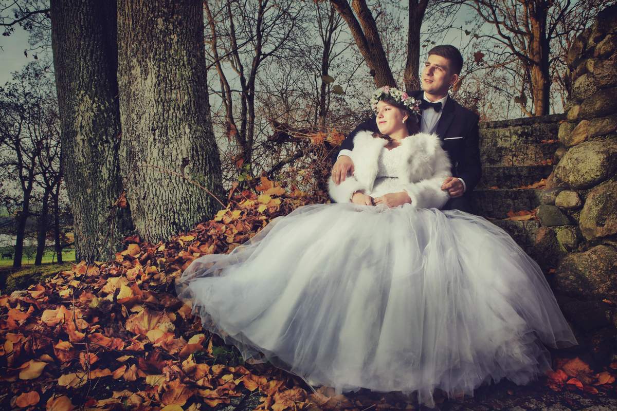 Para młoda siedzi na kamiennych schodach w jesiennym lesie: panna młoda w białej sukni z tiulu i futrzanym bolerku, z wiankiem na głowie, obok pan młody w czarnym smokingu.