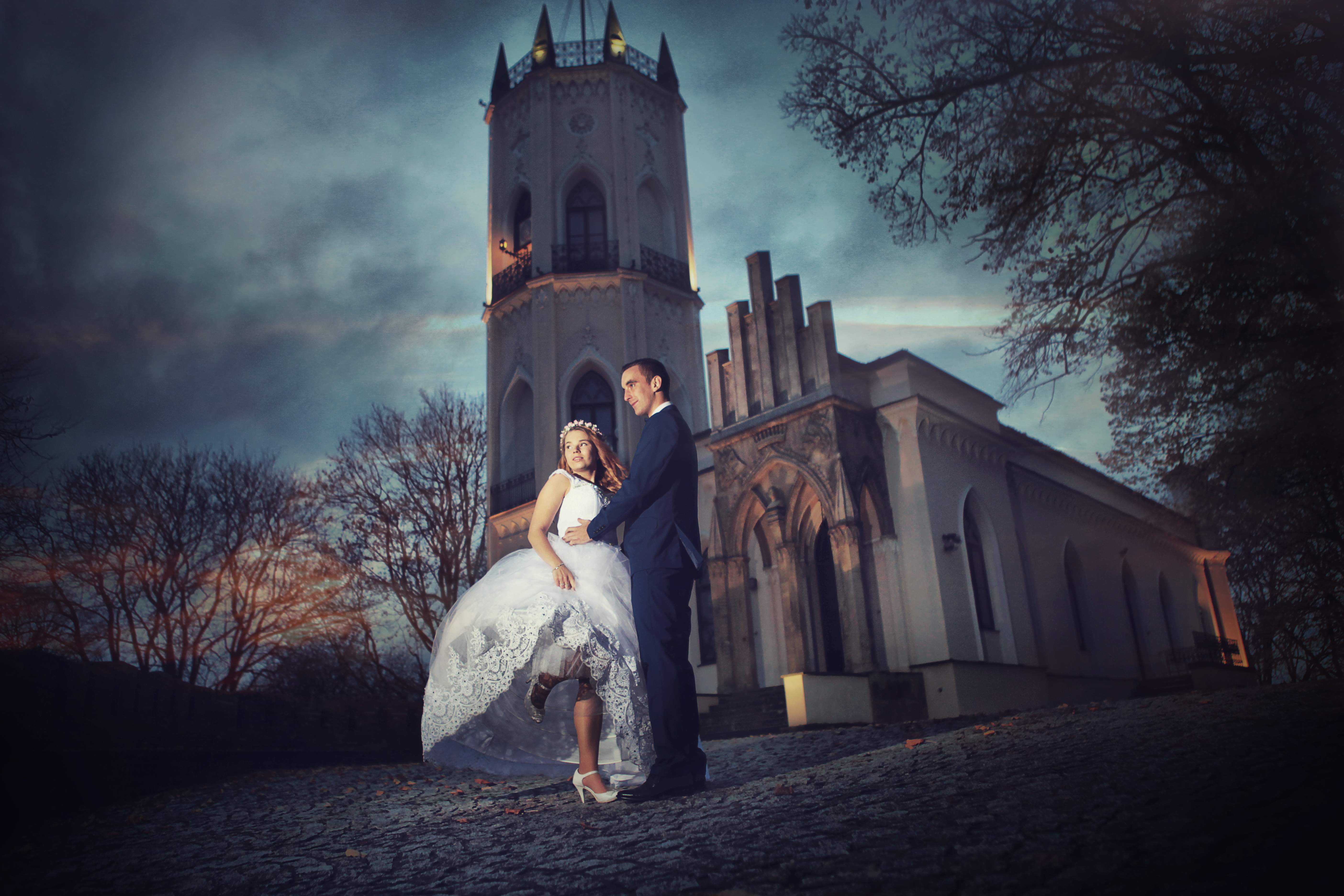 Para młoda w objęciach na tle iluminowanej, gotyckiej architektury z wieżą, na brukowanym placu w pochmurny wieczór.