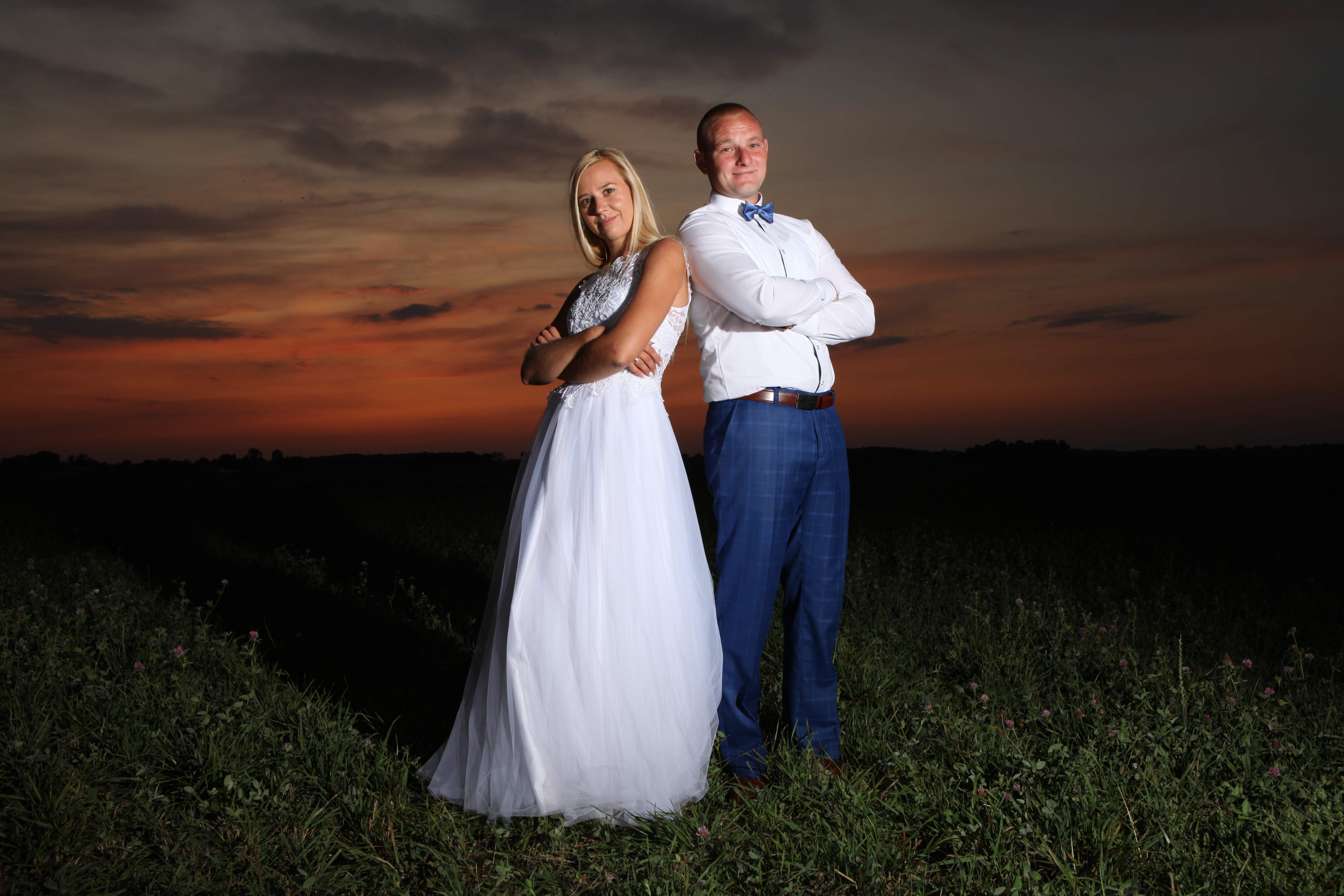 Para młoda, panna młoda w białej sukni i pan młody w niebieskich spodniach i muszce, stojący tyłem do siebie na łące o zachodzie słońca.