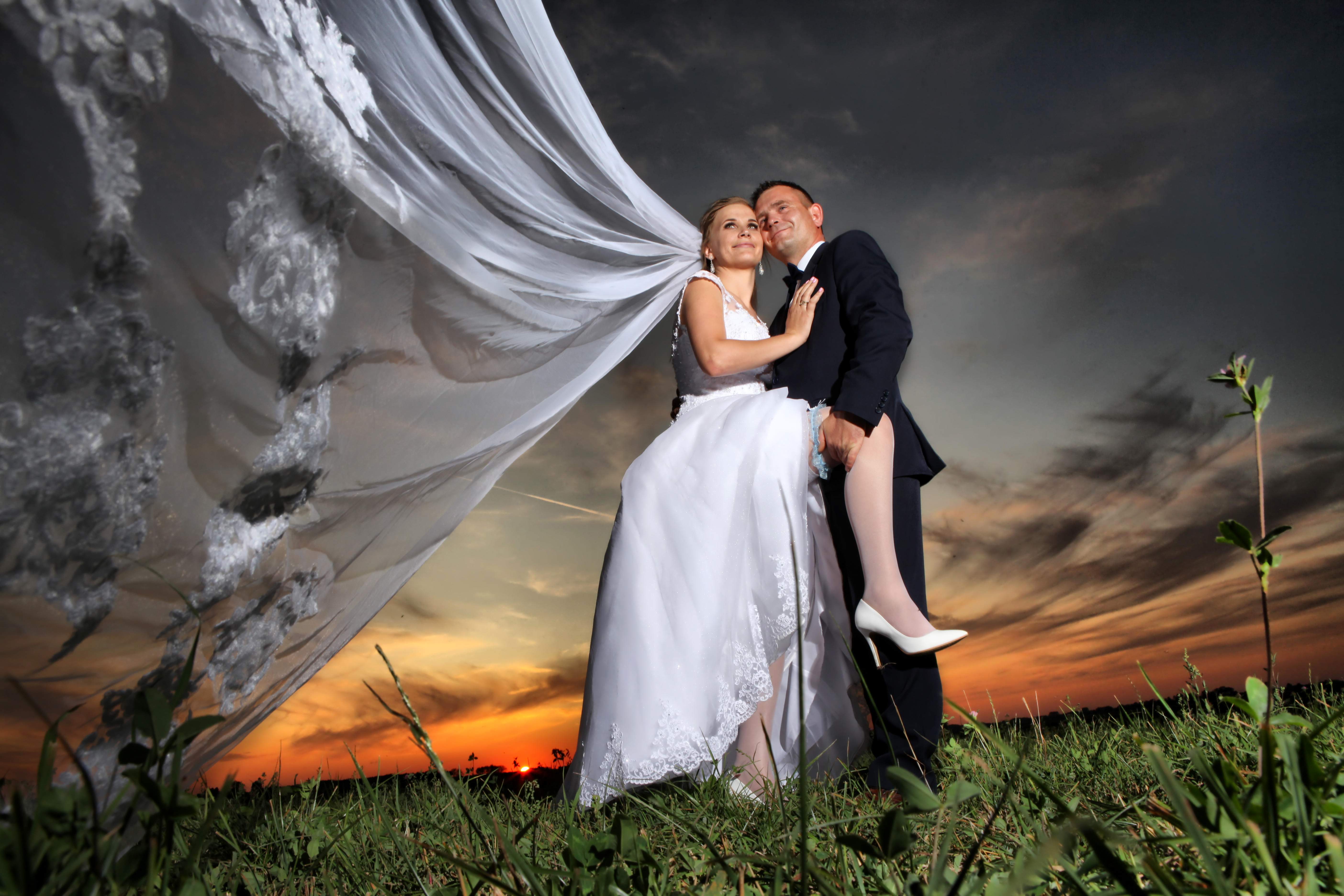 Para młoda na łące podczas zachodu słońca, panna młoda w białej sukni z koronkowym welonem, pan młody w ciemnym garniturze trzyma pannę młodą na rękach, widok z dołu na tle dramatycznego nieba.