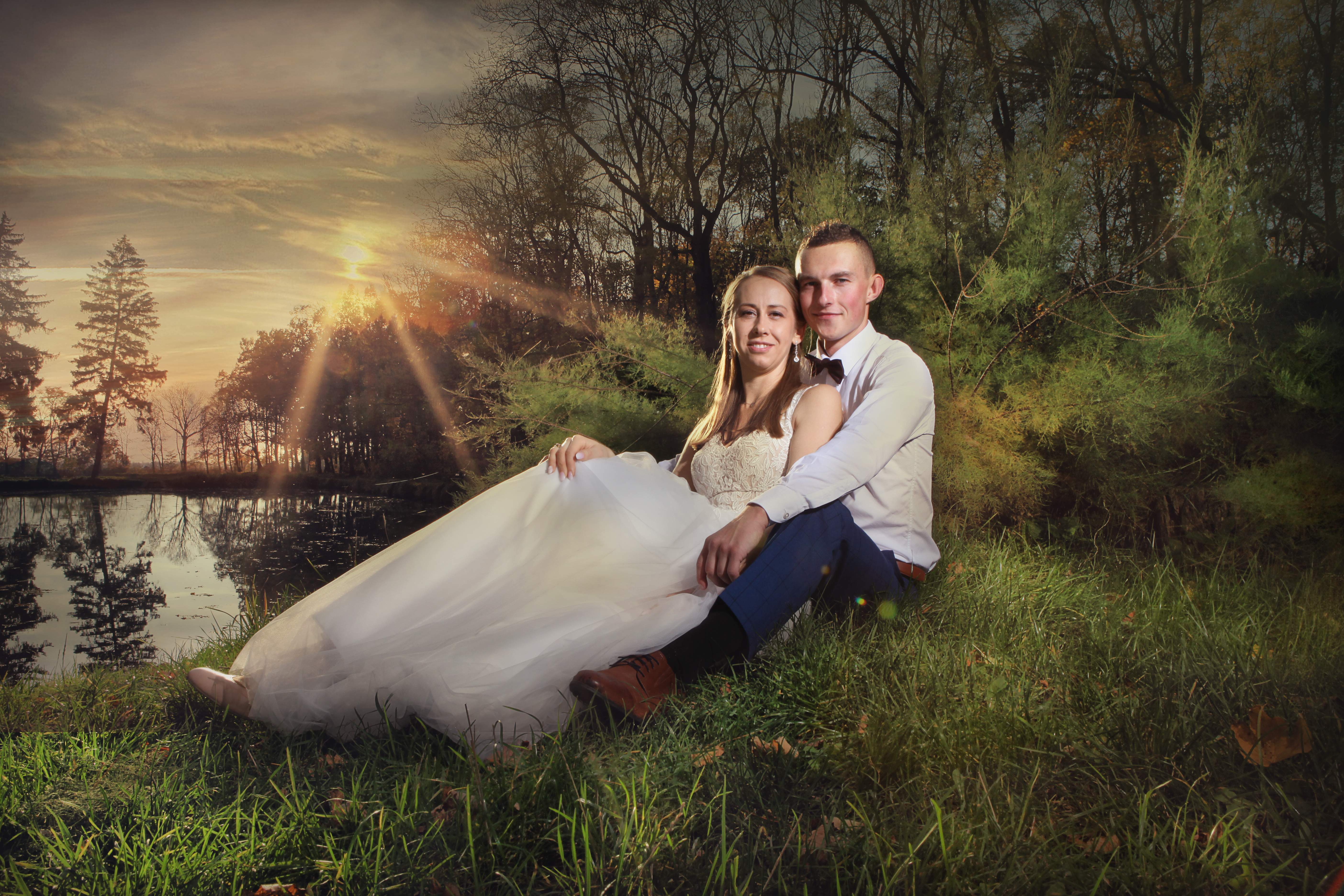 Para młoda siedzi na trawie nad brzegiem stawu o zachodzie słońca. Panna młoda w koronkowej sukni z tiulowym dołem, pan młody w białej koszuli, granatowych spodniach i brązowych butach.