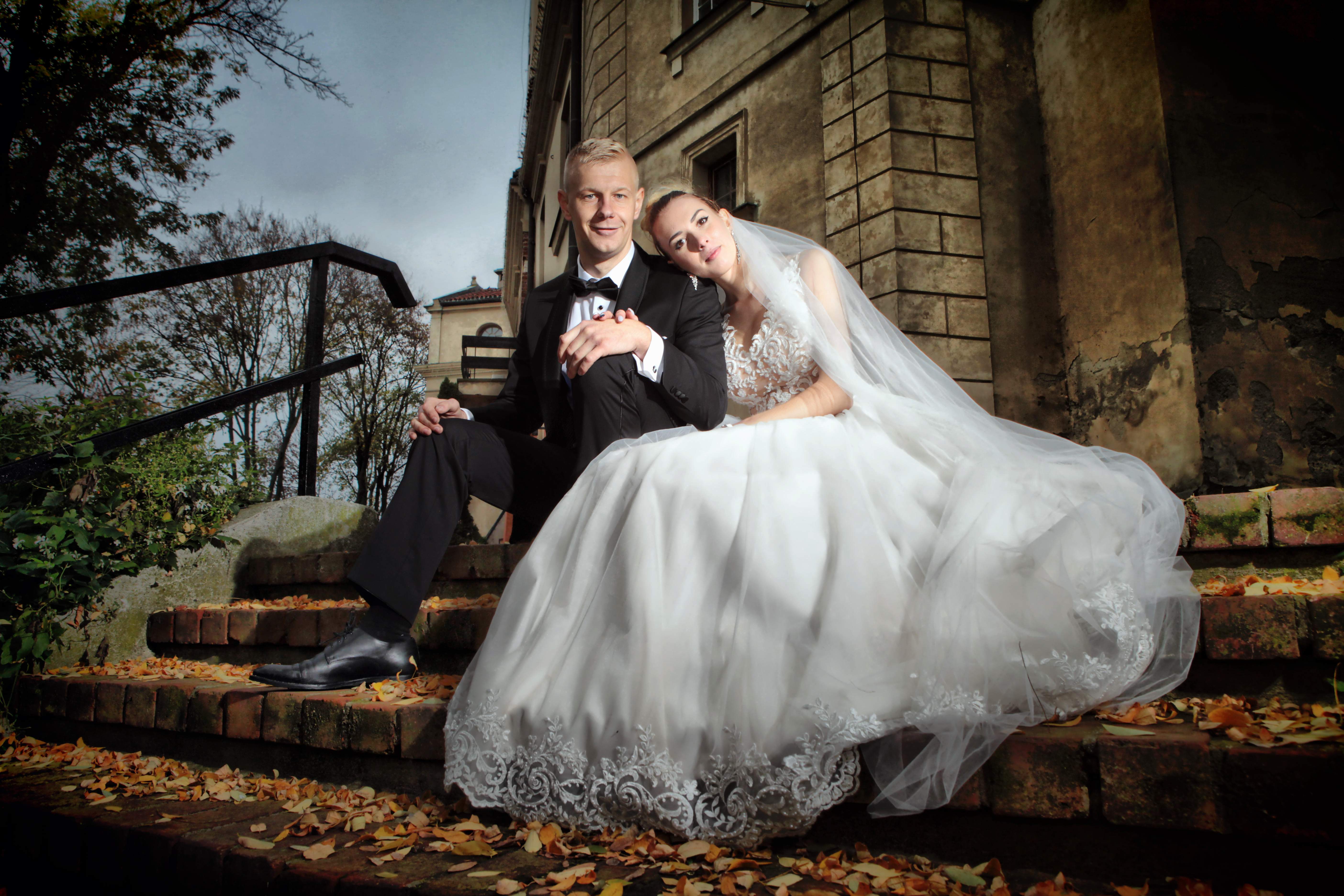 Para młoda siedzi na ceglanych schodach z opadłymi liśćmi, na tle starego budynku z kamiennymi detalami; panna młoda opiera głowę o ramię pana młodego.