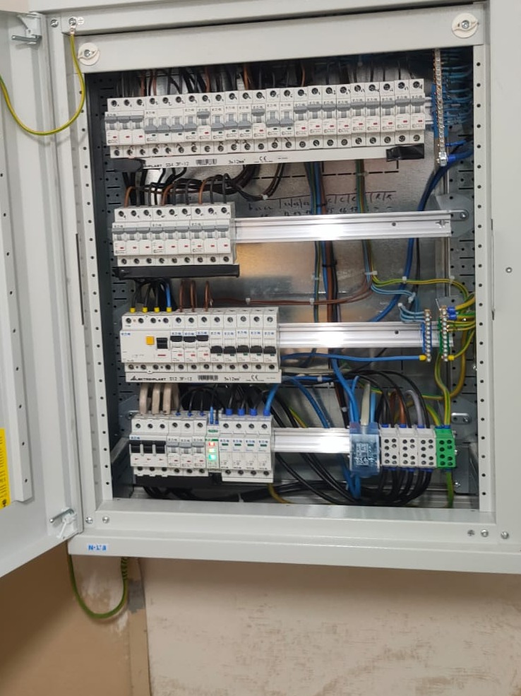 Otwarta szafa rozdzielcza z zamontowanymi bezpiecznikami, przewodami i listwami zaciskowymi, widoczne oznaczenia na obudowie i elementach.