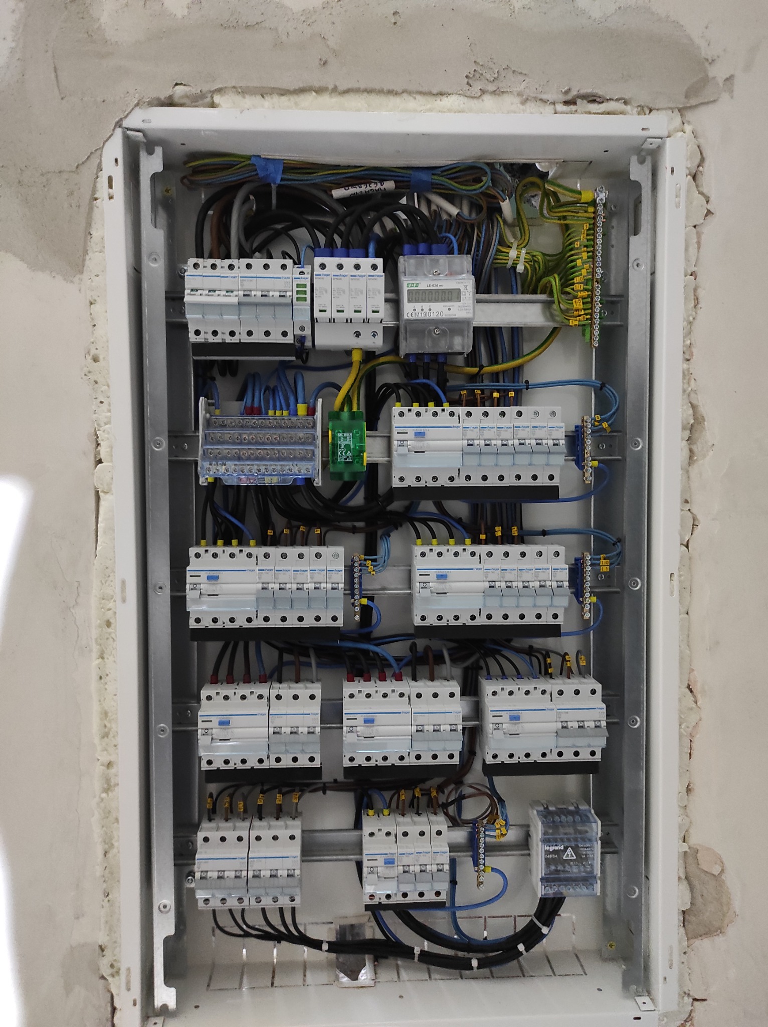Otwarta skrzynka rozdzielcza z zainstalowanymi bezpiecznikami Hager, licznikiem energii elektrycznej LE-02d i przewodami, zamontowana w ścianie w trakcie wykończenia.