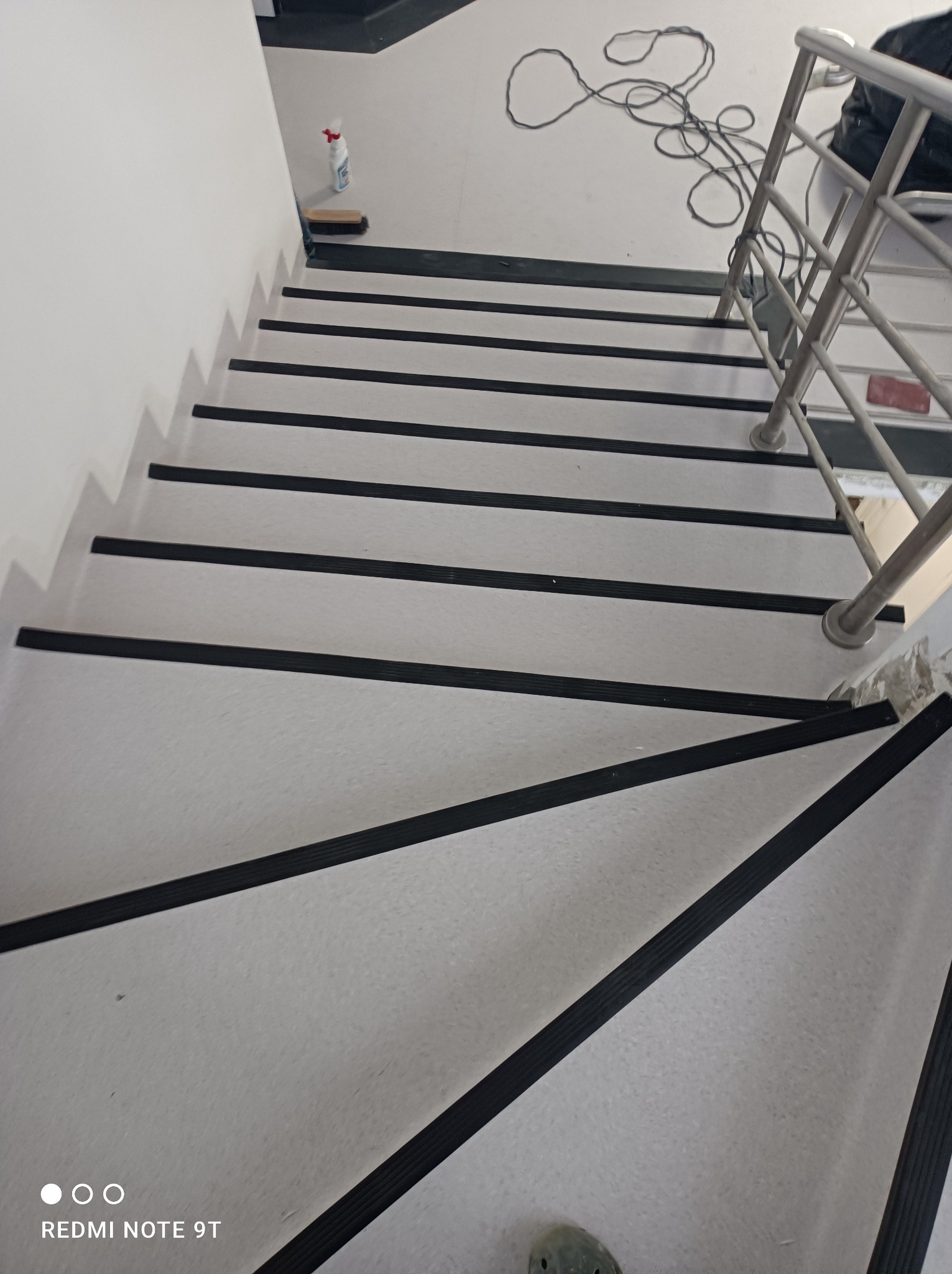 Schody wyłożone jasną wykładziną z antypoślizgowymi, czarnymi paskami, widok z góry na zakręcające schody z metalową barierką.