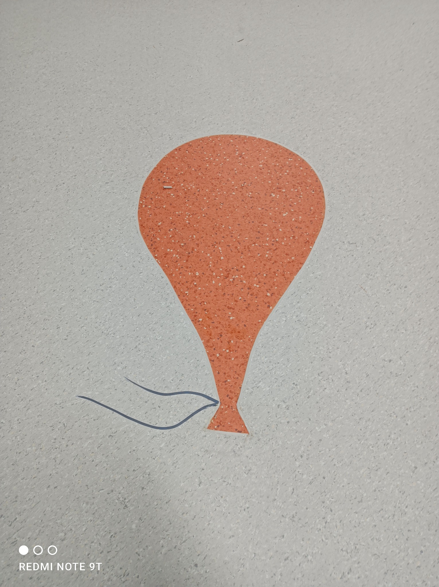 Pomarańczowy wzór przypominający balon na jasnoszarej podłodze z drobnymi plamkami, imitujący wstawkę w podłodze.