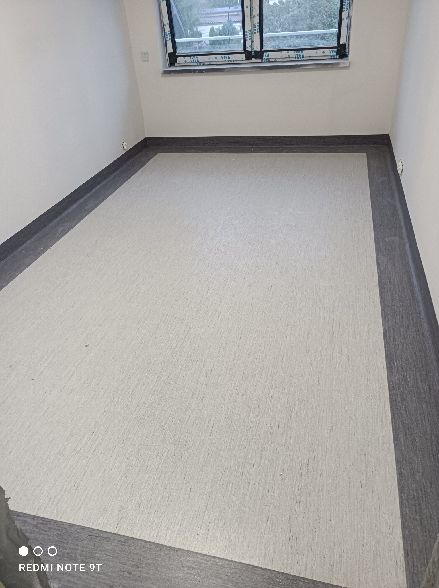 Jasnoszara podłoga z ciemnoszarą listwą przypodłogową w pustym, białym pomieszczeniu z oknem. Widoczny fragment okna z naklejkami producenta szyb.