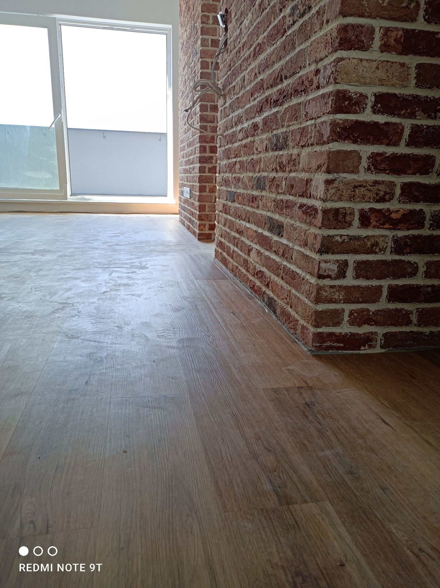 Nowa podłoga z paneli imitujących drewno, świeżo ułożona w pomieszczeniu z ceglaną ścianą i dużym oknem balkonowym.