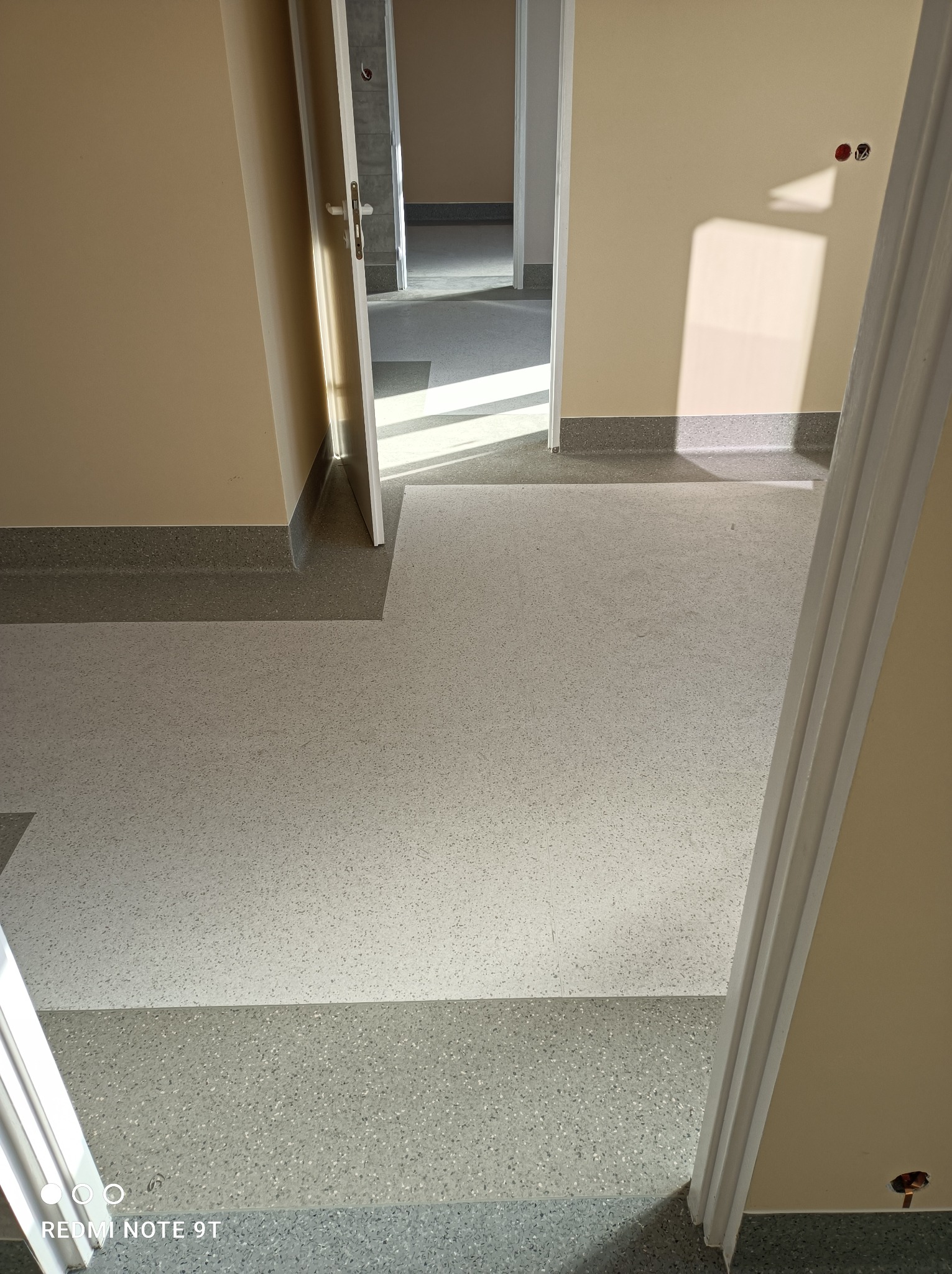 Jasnoszara wykładzina heterogeniczna w korytarzu biurowym, widok przez otwarte drzwi z widocznymi listwami przypodłogowymi i gniazdkami elektrycznymi na ścianie.