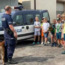 Grupa dzieci obserwuje policjanta z psem policyjnym przy radiowozie podczas pokazu.