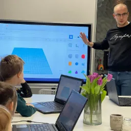 Lekcja projektowania 3D dla dzieci: nauczyciel prezentuje model na ekranie interaktywnym, uczniowie pracują na laptopach, na stole wazon z tulipanami.
