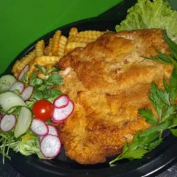 Duży, panierowany kotlet schabowy na czarnym talerzu, podany z frytkami karbowanymi, sałatką z rzodkiewki, ogórka i pomidora, udekorowany zieloną sałatą i natką pietruszki.