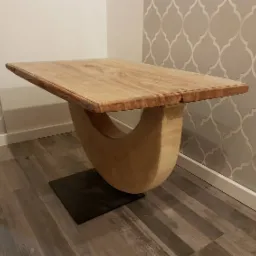 Stolik kawowy z blatem z jasnego drewna i nietypową podstawą w kształcie litery U, umieszczony na czarnej, kwadratowej podstawie, na tle ściany z wzorzystą tapetą.