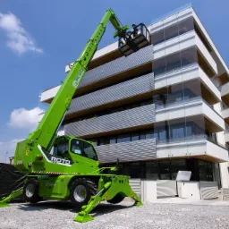 Zielona ładowarka teleskopowa Merlo Roto 40.26 z wysuniętym ramieniem i koszem na tle nowoczesnego budynku z szklanymi balkonami.