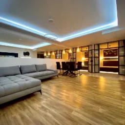 Jasne wnętrze po remoncie z szarą sofą, drewnianą podłogą, oświetleniem LED na suficie i przeszkloną ścianą działową.