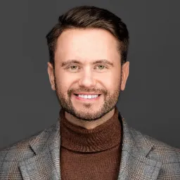 Portret uśmiechniętego mężczyzny w brązowym golfie i szarej marynarce w kratę na jednolitym szarym tle.