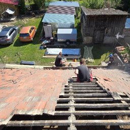 Solid-Dach - Demontaż starej dachówki na drewnianej konstrukcji dachu podczas wymiany, widoczni dekarze w trakcie pracy, w tle zabudowania gospodarcze i samochody.