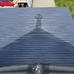 Nowy, blaszany dach w kolorze grafitowym, z widocznym cieniem osoby robiącej zdjęcie, z boku widoczny zadbany trawnik i czerwony samochód.