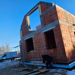 Solid-Dach - Surowy stan budynku z czerwonej cegły, widoczna izolacja fundamentów i pracownik w ciemnej kurtce na tle zimowego krajobrazu i błękitnego nieba.