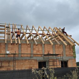 Solid-Dach - Budowa domu jednorodzinnego w trakcie montażu drewnianej konstrukcji dachu, widoczni dwaj pracownicy na wysokościach, jeden z piłą łańcuchową, rusztowania i materiały budowlane na pierwszym planie,...