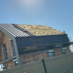 Wymiana dachu w trakcie: widok na dom w budowie z częściowo zdjętym starym pokryciem i zamontowaną blachą na części dachu, rusztowania dookoła budynku.