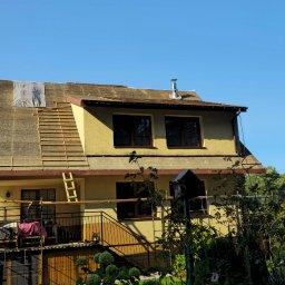 Solid-Dach - Dom w trakcie remontu dachu, widoczna część starego pokrycia, łaty pod nowe pokrycie, komin i drabina oparta o ścianę.