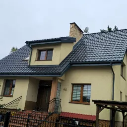 Dom jednorodzinny z nowym, błyszczącym dachem z dachówki ceramicznej w kolorze grafitowym i odnowioną elewacją w odcieniach beżu, widoczny zza czarnego, metalowego ogrodzenia.