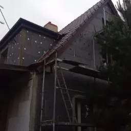 Dom w trakcie ocieplania, widoczne rusztowania, izolacja termiczna na ścianach, fragment dachu z dachówką, osoba z psem na pierwszym planie.