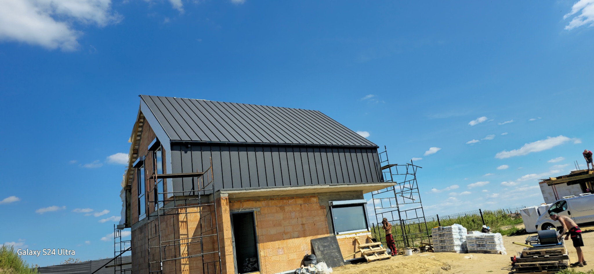 Dom w trakcie budowy z nowym, ciemnoszarym dachem z blachy na tle błękitnego nieba z chmurami. Widoczne rusztowania, materiały budowlane i pracownicy.