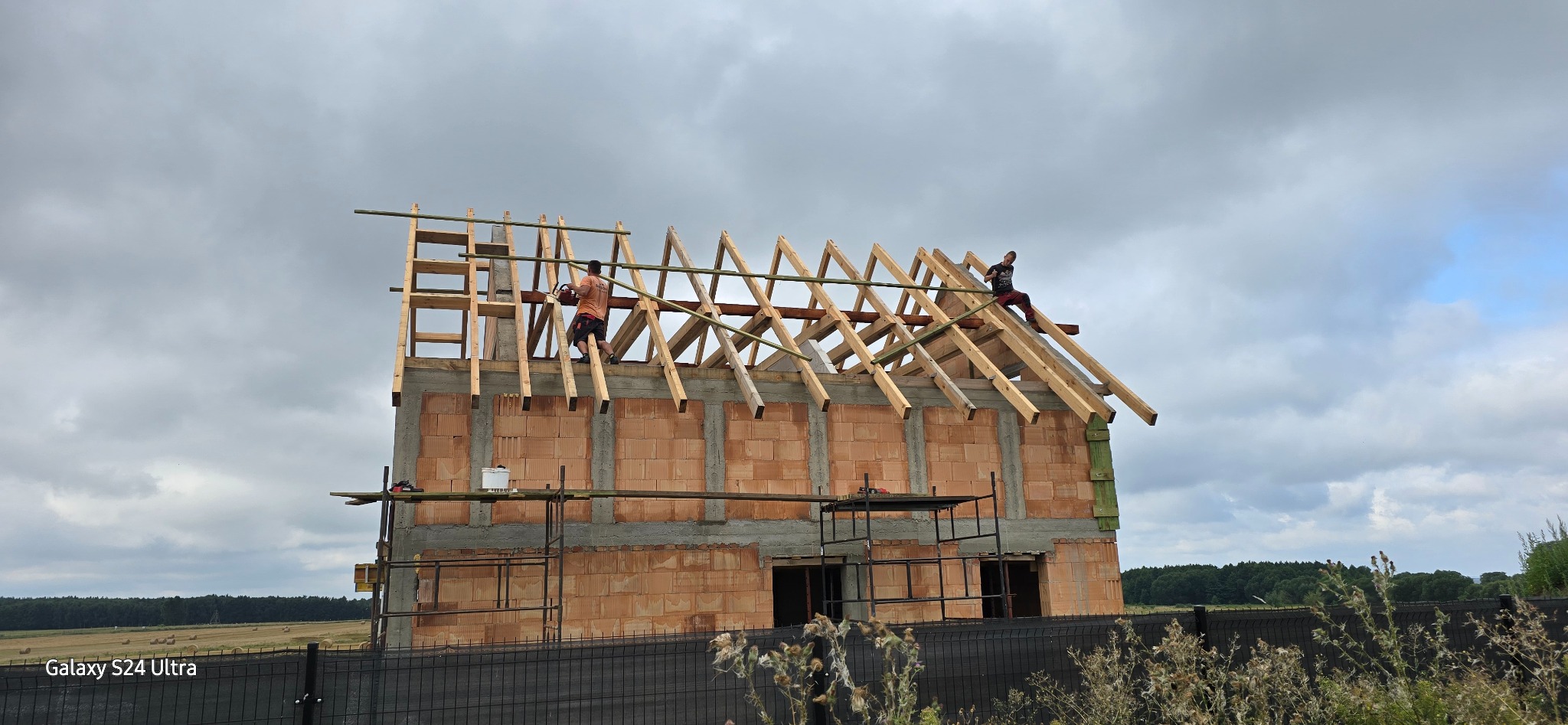 Budowa domu jednorodzinnego w trakcie montażu drewnianej konstrukcji dachu, widoczni dwaj pracownicy na wysokościach, jeden z piłą łańcuchową, rusztowania i materiały budowlane na pierwszym planie,...