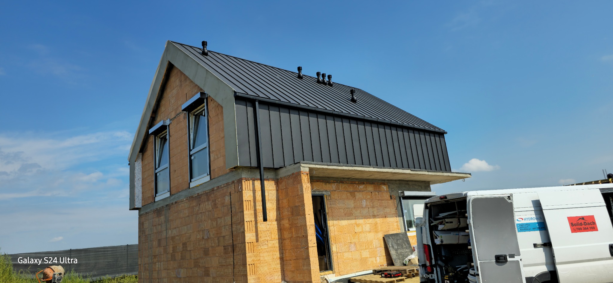 Dom w trakcie budowy z nowym, czarnym dachem z blachy na rąbek stojący, widoczne okna z roletami zewnętrznymi i częściowo wykończona elewacja z cegły ceramicznej, na pierwszym planie samochód...