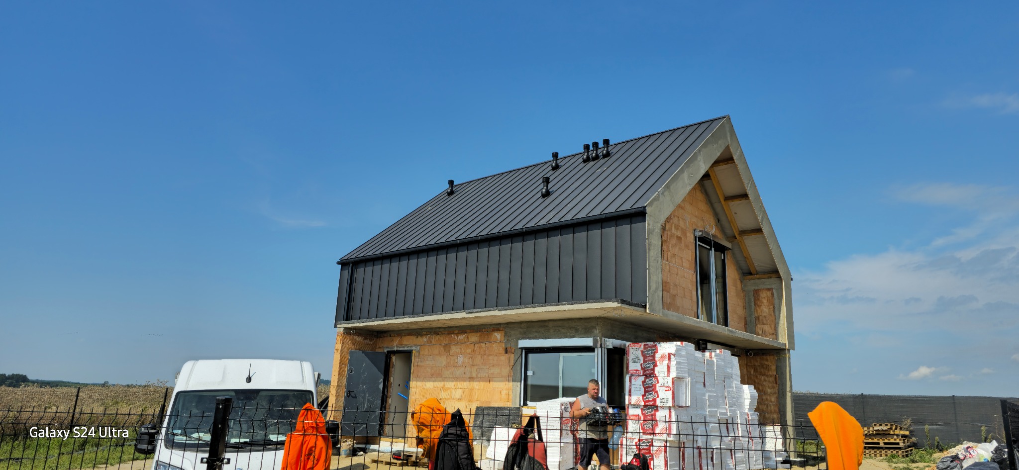 Nowoczesny dom w trakcie budowy z ciemnym, metalowym dachem, widoczne materiały izolacyjne, pracownik i samochód dostawczy na tle błękitnego nieba.