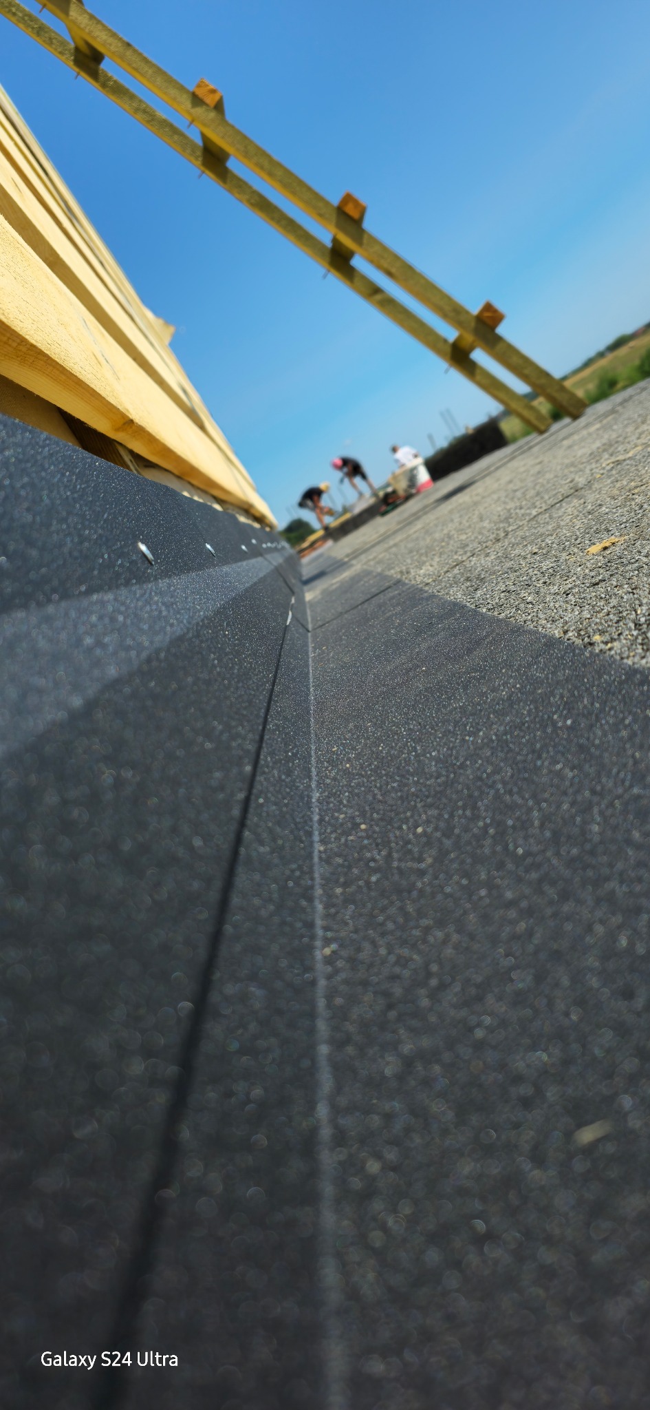 Czarny, teksturowany materiał dachowy z widoczną krawędzią i mocowaniami, w tle ekipa montująca na drewnianej konstrukcji dachu pod błękitnym niebem.