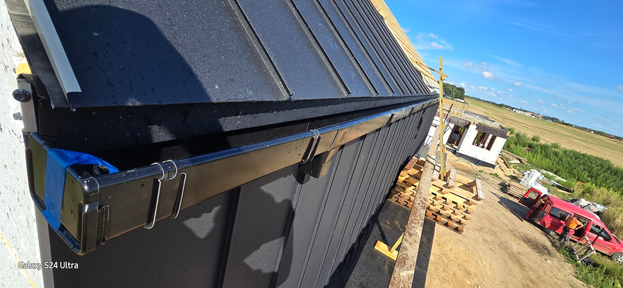 Czarny, metalowy dach z zamontowaną rynną, zabezpieczoną niebieską taśmą, widok z góry na budowę domu i czerwoną furgonetkę w tle.