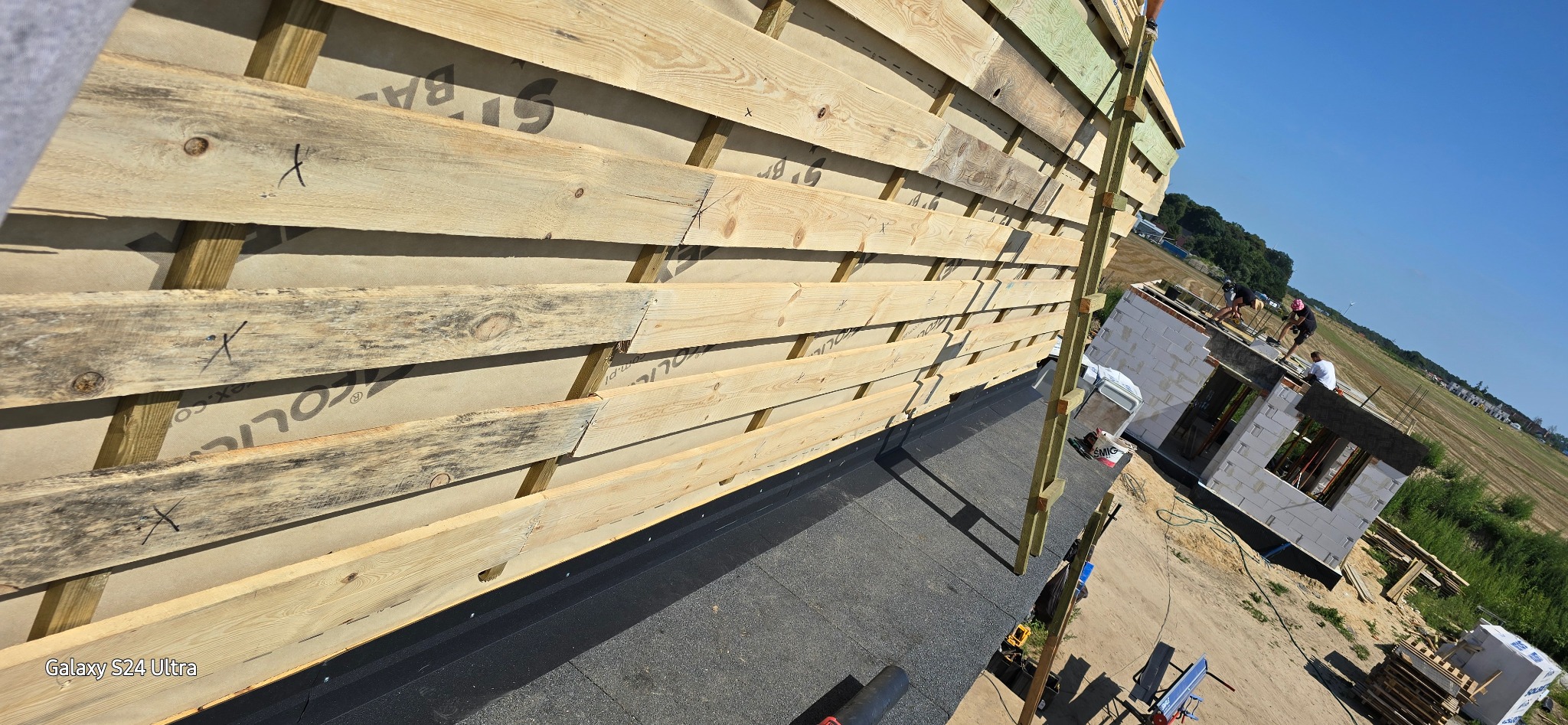 Montaż drewnianej konstrukcji dachu na folii izolacyjnej, widoczne elementy więźby dachowej i fragment budynku w trakcie budowy w tle.