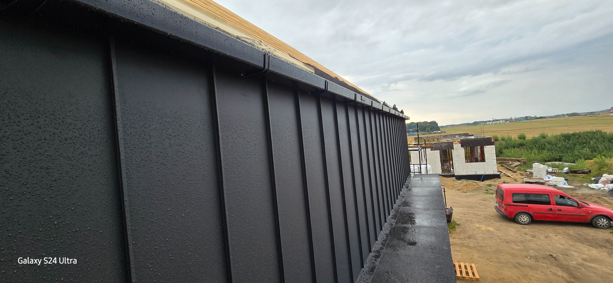 Czarny, blaszany dach na mokro, widoczne krople deszczu, w tle budowa domu z czerwonym samochodem dostawczym w pobliżu.
