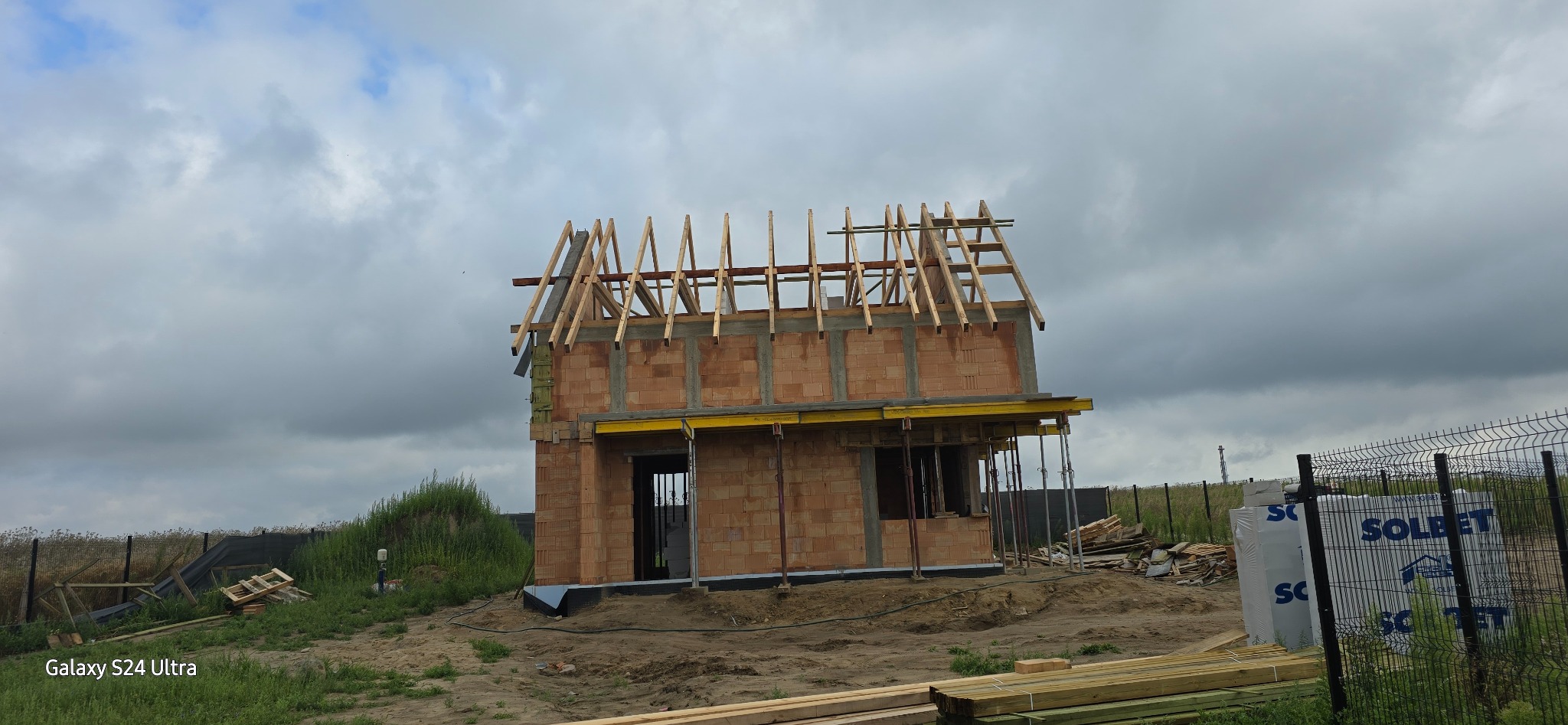 Budowa domu jednorodzinnego w stanie surowym otwartym: murowane ściany z pustaków ceramicznych, częściowo wykonana więźba dachowa z drewnianych krokwi, szalunki stropowe podtrzymywane przez stemple...