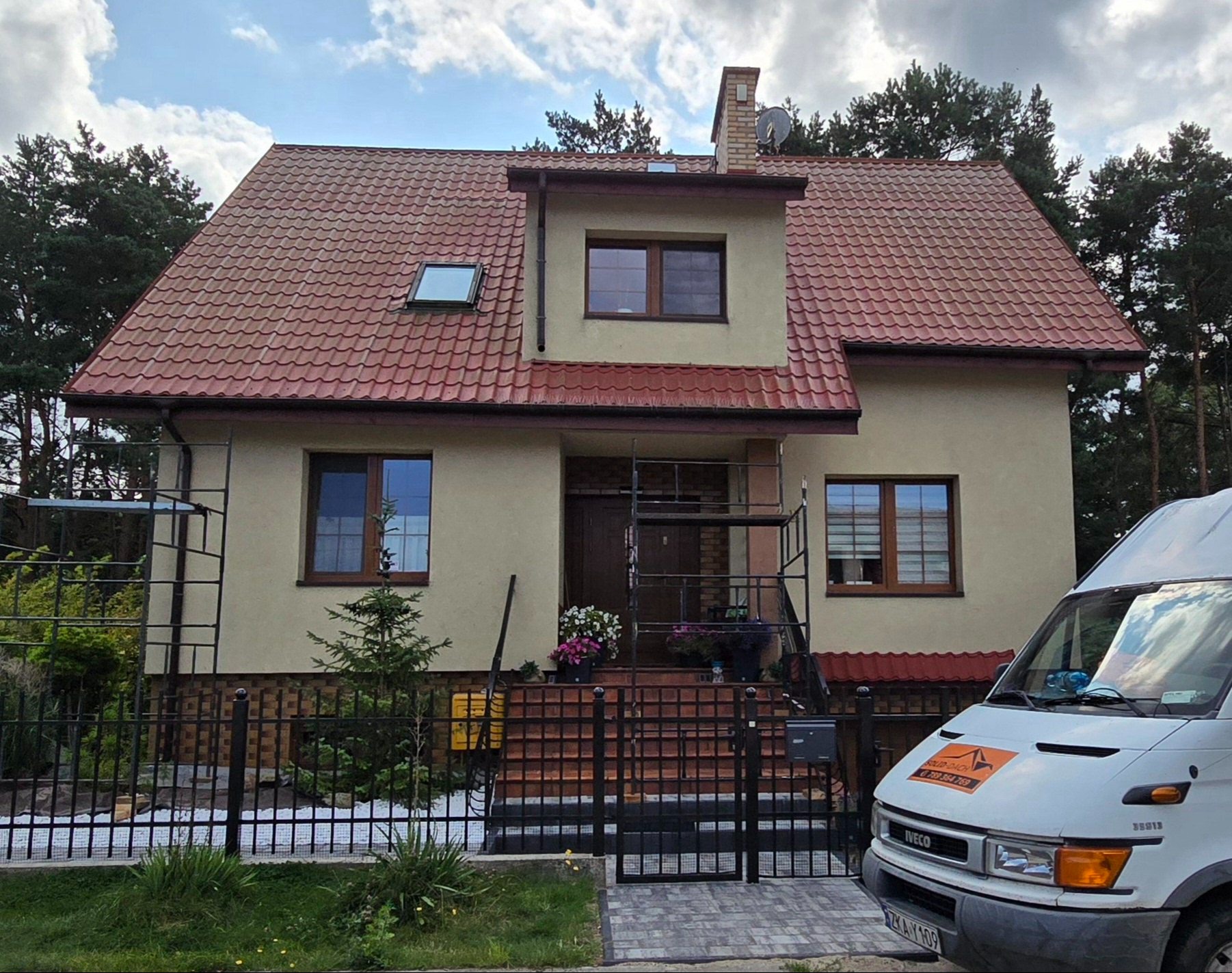 Dom jednorodzinny z nowym, czerwonym dachem z dachówki, widoczne rusztowanie przy wejściu i firmowy samochód dostawczy.