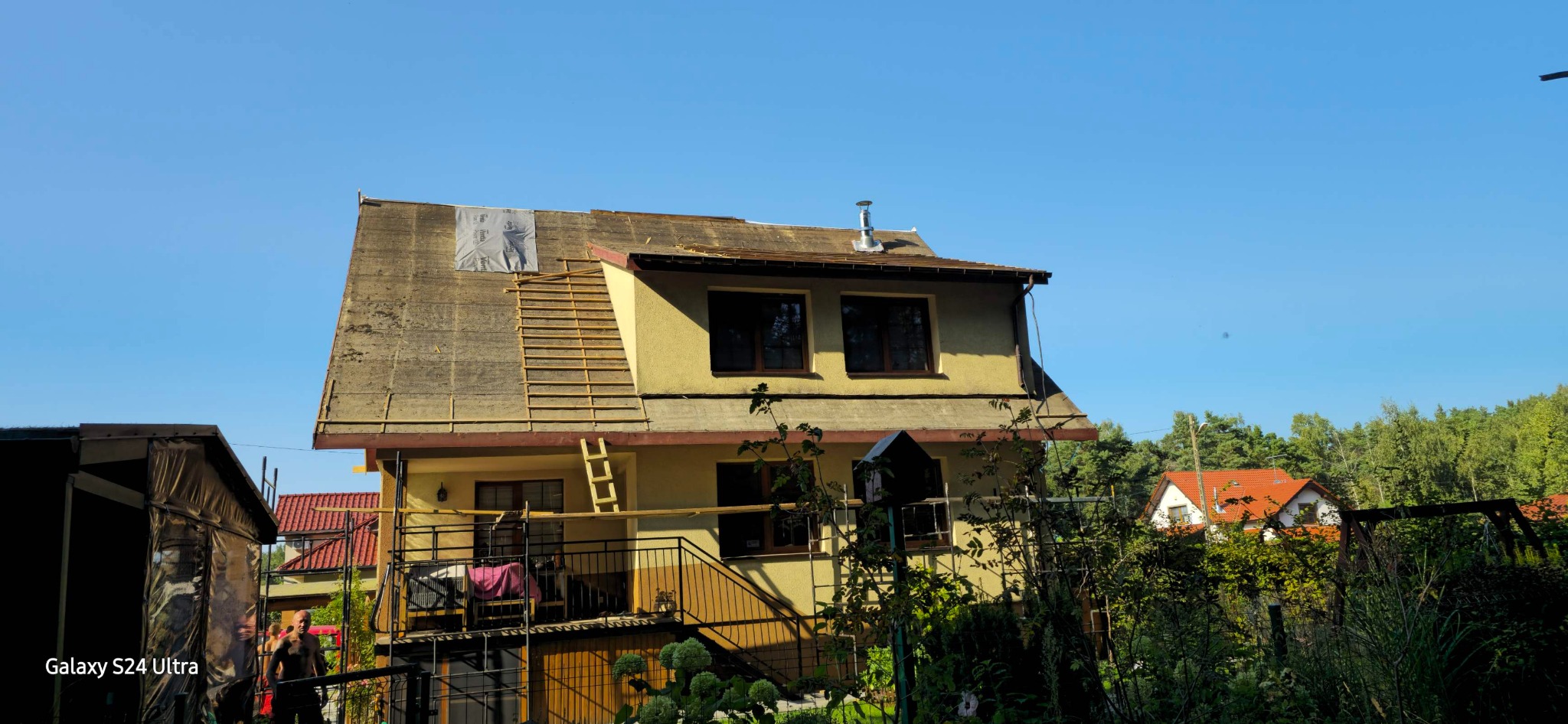 Dom w trakcie remontu dachu, widoczna część starego pokrycia, łaty pod nowe pokrycie, komin i drabina oparta o ścianę.